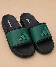 A pair of Adidas slide sandals with black footbeds and straps, featuring a textured green strap with a green Adidas logo, set against a solid beige background.