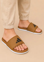 A person wearing tan slide sandals with a black Adidas logo and white soles, paired with beige pants, against a solid beige background.