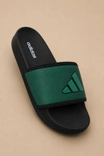 An Adidas slide sandal with a black footbed and sole, featuring a wide textured green strap with a green Adidas logo, set against a solid beige background.