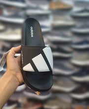 A hand holds an Adidas slide sandal with a black and white design, set against a blurred background of shelves filled with shoes.