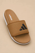 An Adidas slide sandal in a tan colorway, featuring a black three-bar logo on the strap, a white outsole, and the Adidas wordmark printed on the footbed, set against a solid beige background.