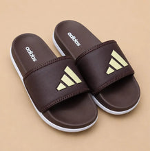A pair of Adidas slide sandals in dark brown with a textured upper strap featuring the brand's signature three stripes in a pale yellow-green, set against a solid beige background.