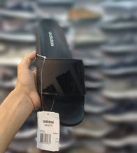 A hand holds an Adidas Adilette slide sandal in black, showing the side profile with the brand name and a white product tag. The background is a highly blurred, out-of-focus display of many shoes.