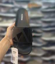 A hand holds an Adidas slide sandal with a white retail tag, set against a blurred background of shelves filled with shoes.