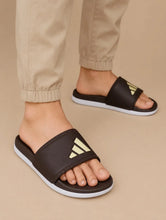 A person wearing dark brown slide sandals with a gold Adidas logo and white soles, paired with beige pants, against a solid beige background.