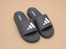 A pair of Adidas slide sandals in dark gray with white logo details, displayed on a solid beige background.