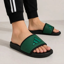 A person wearing green and black slide sandals with the Adidas three-stripes logo on the strap, shown from the feet up against a light gray studio background.