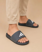 A person wearing dark gray Adidas slide sandals with beige jogger pants, shown from the calves down against a plain beige studio background.