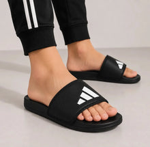 A person wearing black Adidas slide sandals with the classic white three-stripes logo, paired with black jogger pants featuring white stripes.