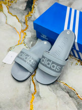A pair of Adidas slide sandals in light gray with a textured logo strap, displayed next to their blue Adidas shoebox on a marble-patterned surface.