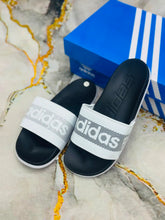 A pair of Adidas slide sandals with black footbeds and white straps featuring the Adidas logo, displayed next to a blue Adidas shoebox on a marble-patterned surface.
