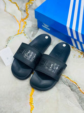 A pair of black Adidas slide sandals with embossed branding on the straps, displayed next to a blue Adidas shoebox on a light marble-patterned surface.