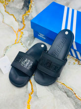 A pair of black Adidas slide sandals with embossed branding on the straps, displayed next to a blue Adidas shoebox on a marble-patterned surface.
