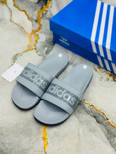 A pair of Adidas slide sandals in gray with a textured logo strap, shown next to their blue Adidas shoebox on a marble-patterned surface.