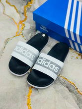 A pair of Adidas slide sandals with white straps and black footbeds, placed next to a blue Adidas shoebox on a marble-patterned surface.