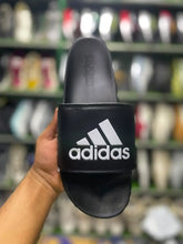 A black Adidas slide sandal with a white logo, held by a hand against a blurred background of shoe shelves.