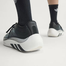 A close-up of a person's feet wearing Adidas sneakers, showing the side and rear of the shoe with black socks on a light gray studio background.