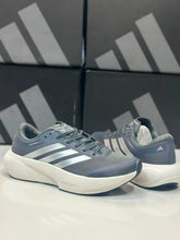 A pair of Charcoal Silver Adidas Supernova Rise 3 in slate gray and white, displayed on a reflective white surface with black Adidas shoeboxes in the background.