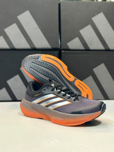 An Dark Grey Orange Gradient Adidas Supernova Rise 3 in dark gray and orange with white stripes, shown alongside its matching orange and gray outsole against a background of black Adidas shoeboxes.