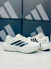 A pair ofWhite Black Adidas Supernova Rise 3  displayed on a reflective white surface with dark Black Adidas shoeboxes in the background.