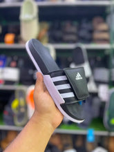 A hand holding an Adidas slide sandal with a black strap featuring three white stripes and a white logo, set against a blurred background of shoe shelves.