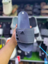 A person's hand holds an Adidas slide sandal in a retail store setting. The sandal features a gray strap with the black Adidas logo and three stripes, and a matching gray footbed.