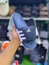 A navy blue Adidas slide sandal with white stripes and a white logo, held by a hand in a shoe store.