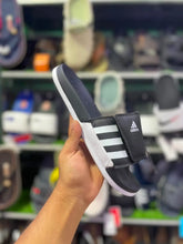 A hand holding an Adidas slide sandal with a black strap, white three-stripes, and a white sole, set against a blurred background of shoe shelves.