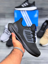 A hand holds a black Adidas Ultraboost sneaker, with other shoes and an Adidas box blurred in the background on a gray wood floor.