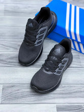 A pair of black Adidas running shoes with gray side stripes, displayed on a gray wood-look floor next to a blue Adidas box.