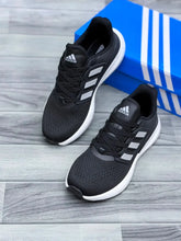 A pair of Core Black White Adidas Ultraboost 5 running shoe with white stripes and white soles, displayed on a gray wood-look floor next to a blue Adidas box.