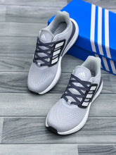 A pair ofLight Grey White Adidas Ultraboost 5 running shoes with dark gray laces, displayed on a gray wood-look floor next to a blue Adidas shoebox.