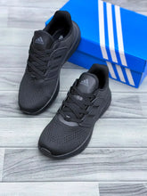A pair of Triple Black Adidas Ultraboost 5 running shoe displayed on a gray wood-look floor next to a blue Adidas shoebox with white stripes.