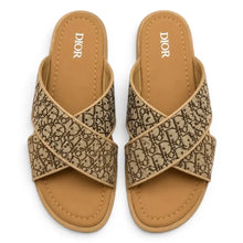 A pair of Dior slide sandals featuring a beige and brown monogram patterned upper with a tan footbed, shown from a top-down view on a white background.