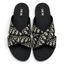 A pair of Dior slide sandals featuring a black and beige monogram pattern across the straps, with black soles and the brand name 'DIOR' printed in white on the insoles, set against a plain white background.