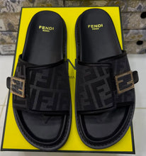 A pair of black Fendi slide sandals with the signature FF monogram pattern and gold-tone buckle, resting on a bright yellow Fendi box.
