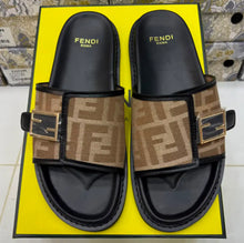 A pair of Fendi slide sandals with the iconic brown monogram canvas straps and gold-tone FF logos, displayed on a bright yellow Fendi box.