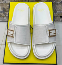 A pair of white Fendi slide sandals with gold-tone buckle details, displayed on top of a bright yellow Fendi shoebox.