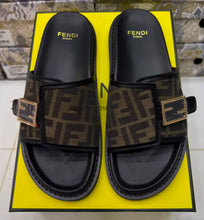 A pair of Fendi Roma slide sandals with the signature brown and black FF monogram canvas straps and gold-tone buckle details, displayed on top of a bright yellow Fendi box.