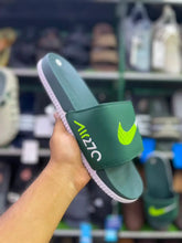 A Nike Air 270 slide sandal in dark green with a neon yellow-green Swoosh and white 'AIR 270' text on the strap, held by a hand in front of a blurred shoe store background.