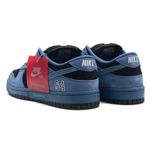 A pair of blue and black Nike x Supreme SB Dunk Low sneakers, featuring a red hangtag with the Nike and Supreme branding, on a white background.